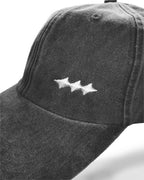 A close-up of a dark gray washed cotton baseball cap featuring a white embroidered logo of three connected stars on the front panel.